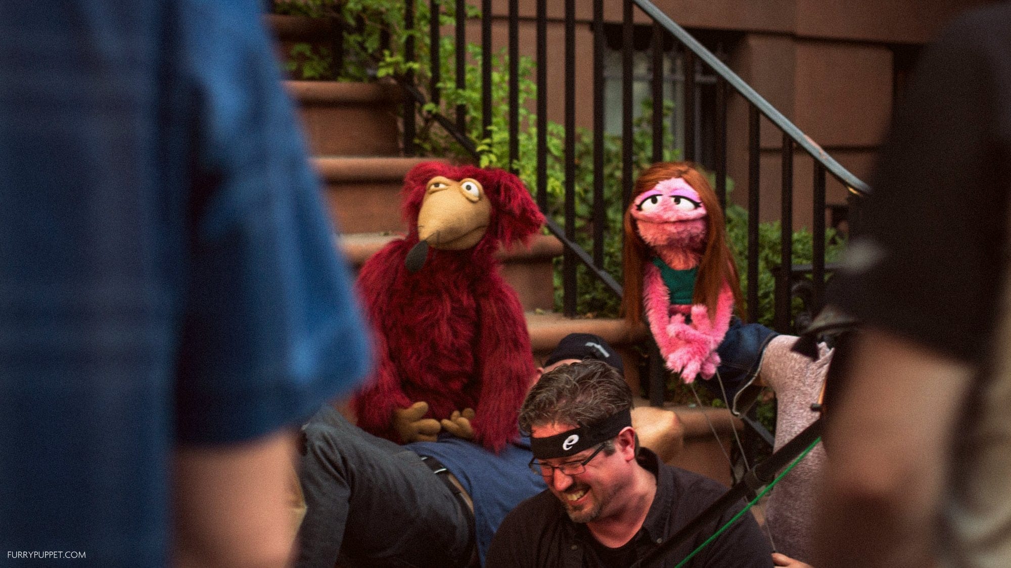 Behind-the-scenes view of a production, with a red furry puppet and a pink puppet with long brown hair posed on a brownstone staircase as crew members operate them.