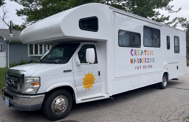 White mobile art center RV parked outside a house, with colorful “Creative Wanderings Art Center” lettering and a cheerful smiling sun logo.