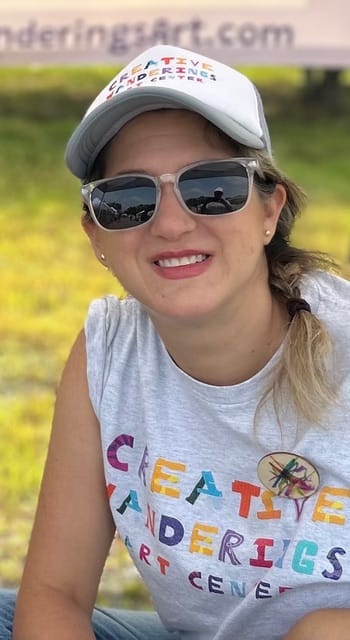 Smiling woman outdoors in mirrored sunglasses and a white cap, wearing a colorful creative-arts T-shirt against a sunny green background.