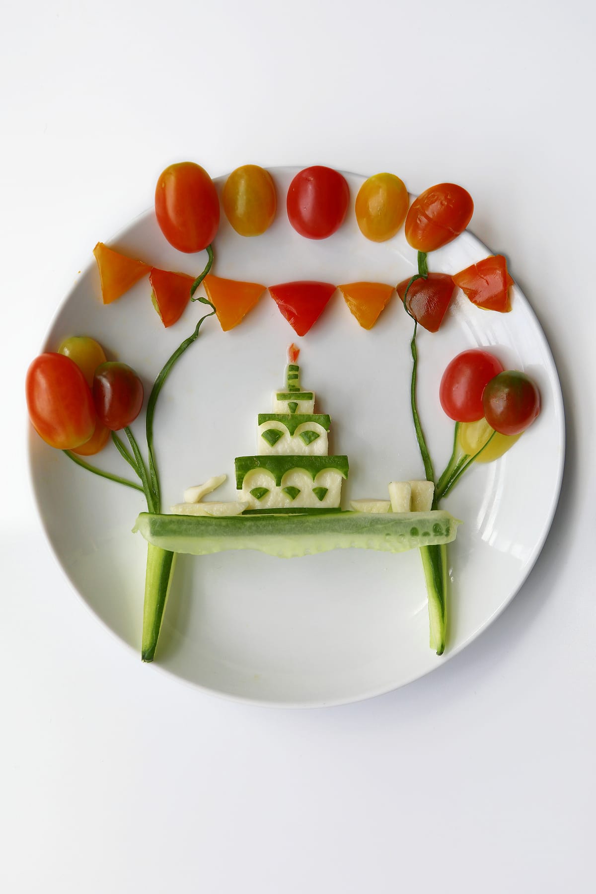 Food art on a white plate depicting a birthday celebration, with a layered cucumber-and-cheese cake, a small candle, tomato balloons, cucumber stems and a string of orange and red vegetable bunting.