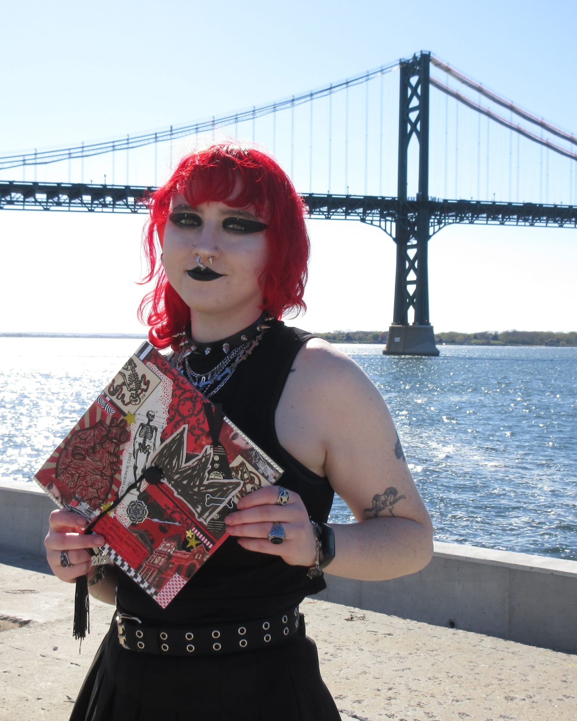 Killian Finn Paris, with bright red hair and bold black makeup, holds up a graduation cap decorated with collage. The cap shows an anatomical heart, a skeleton and red patterns. Behind them, a suspension bridge stretches over sparkling water.