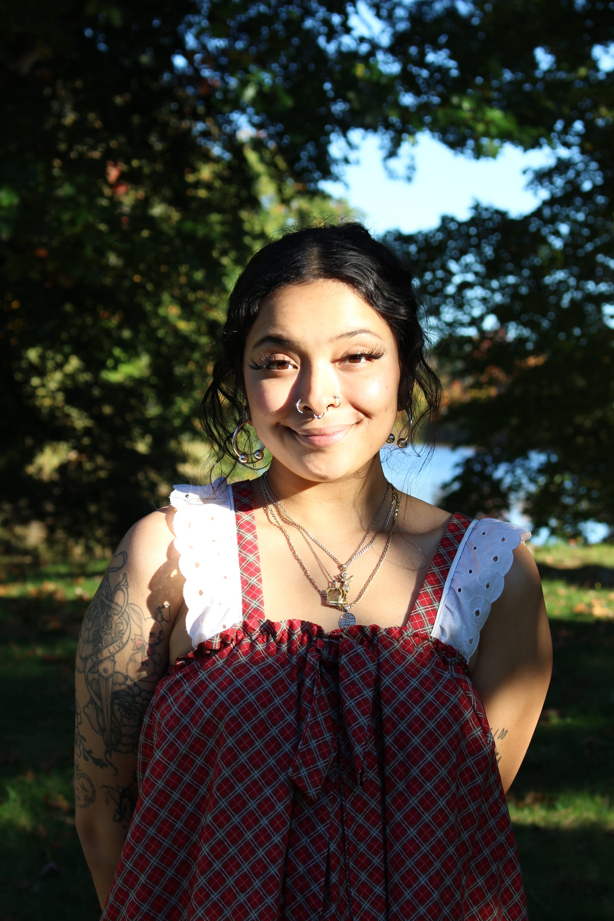Janise Nieves smiles in the sunlight, their dark hair pulled back, wearing nose rings, hoop earrings, layered necklaces and a red plaid top with white eyelet ruffle straps, with tattoos on their arm. Trees and water are behind them.