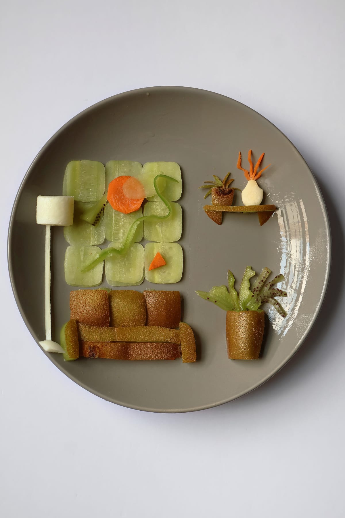 Food art on a gray plate showing a living room made from fruit and vegetable pieces, including a kiwi-slice couch, floor lamp, wall shelf with plants and potted plant.
