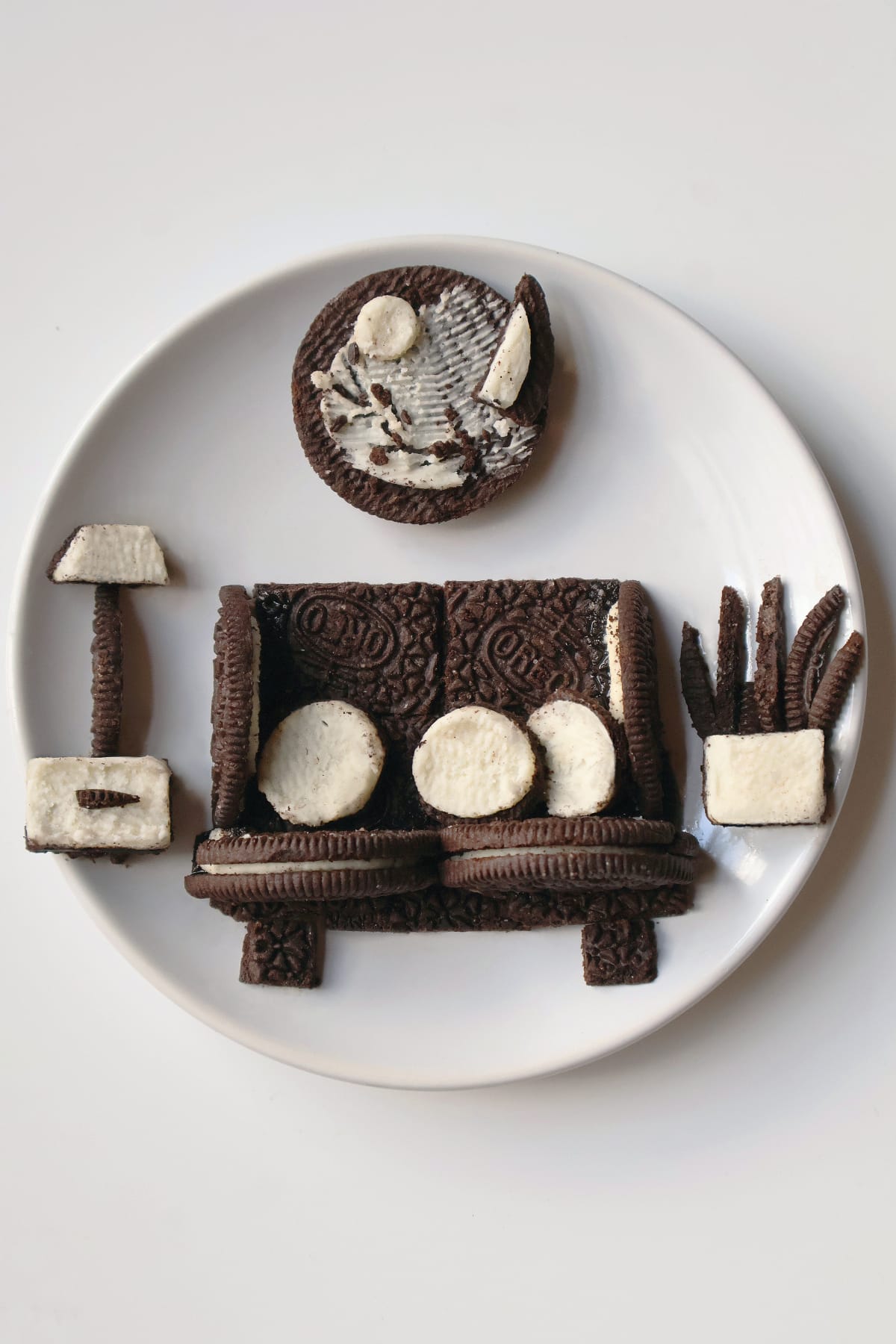 Food art arranged on a white plate to resemble a living room, with an Oreo-cookie couch, a cream-and-cookie television, a tall floor lamp, a potted plant and a round wall decoration.
