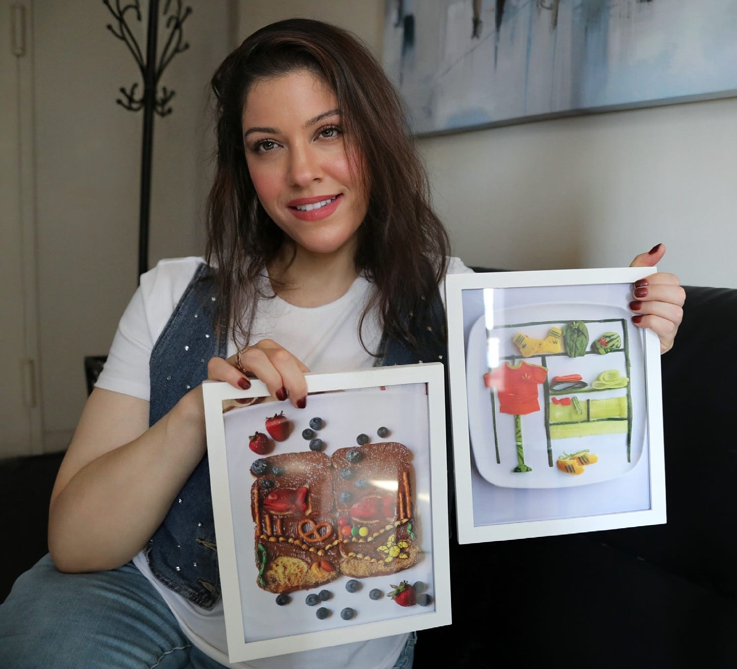 Woman seated on a couch, smiling and holding two white-framed photographs of food art: a toast design decorated with fruit and a clothing display made from colorful food.
