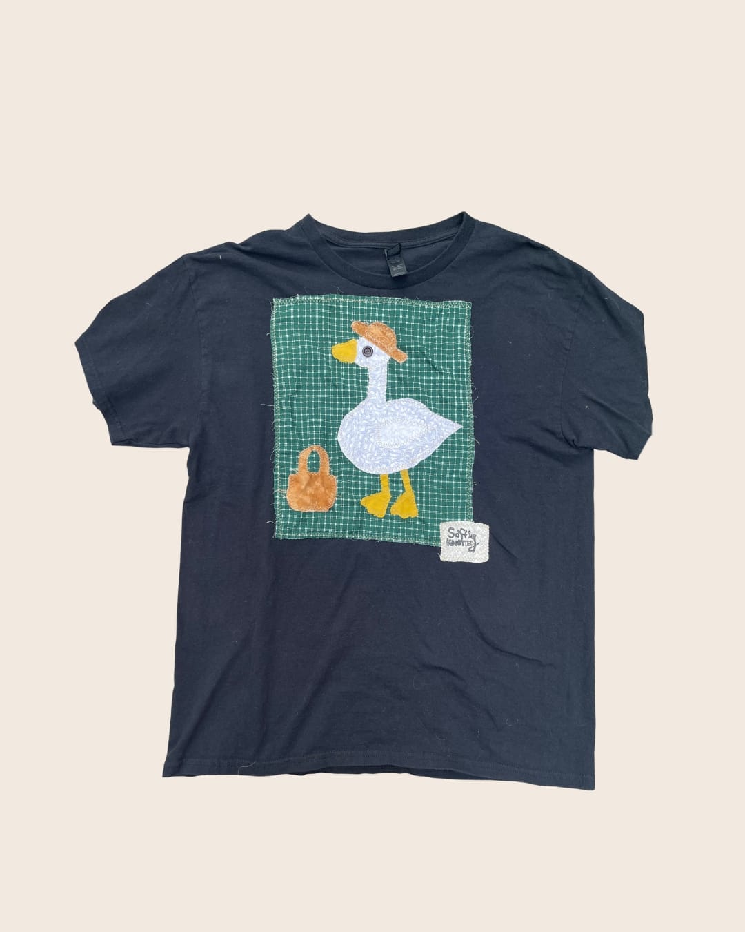 A black T-shirt with a hand-sewn appliqué of a white goose wearing a straw hat beside a small basket, on green gingham fabric, with a “Softly Knotted” patch below.