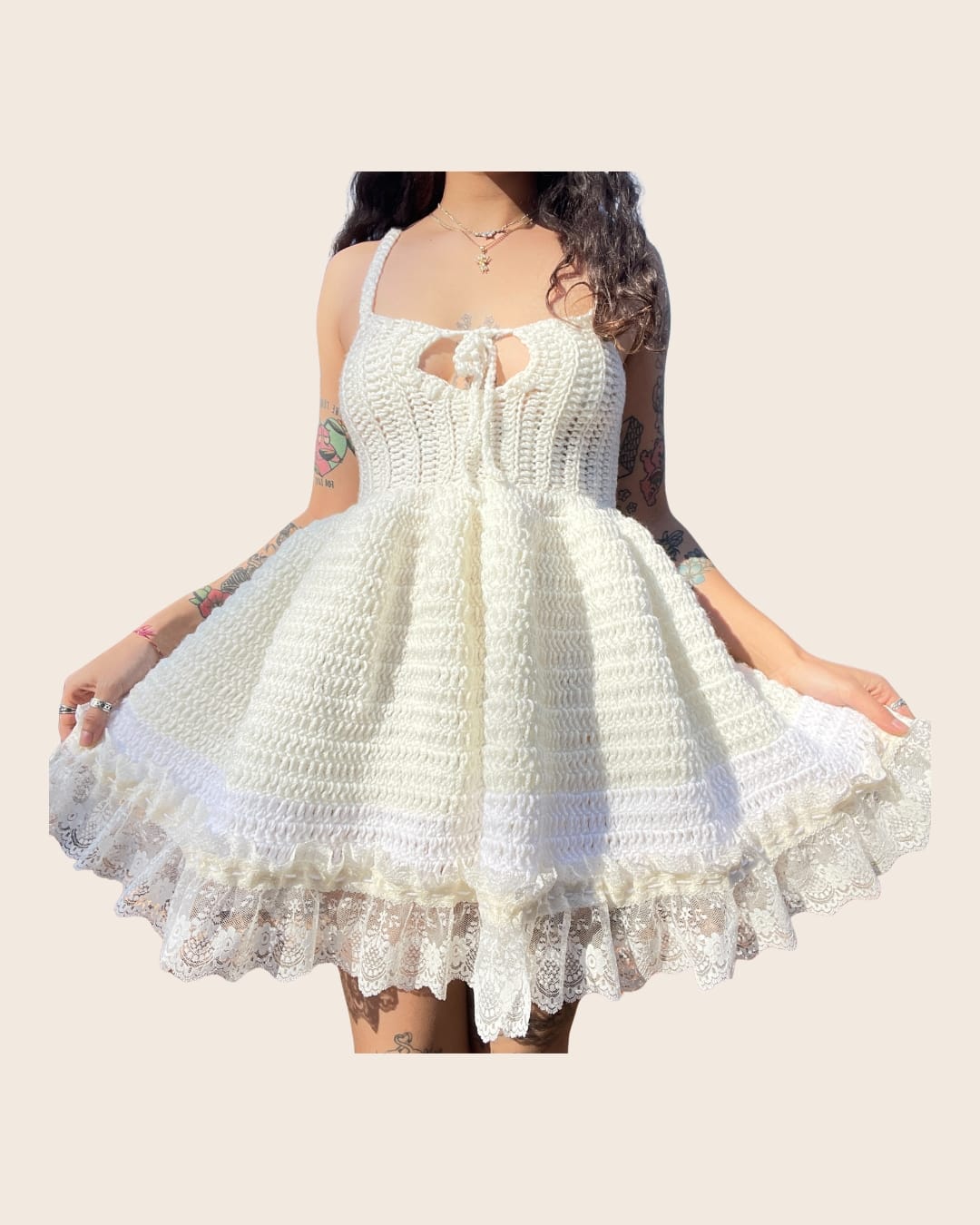 A person holds out the skirt of a cream crochet mini dress with a corset-style bodice, a tie-front keyhole neckline, a full skirt and a white lace ruffle hem.