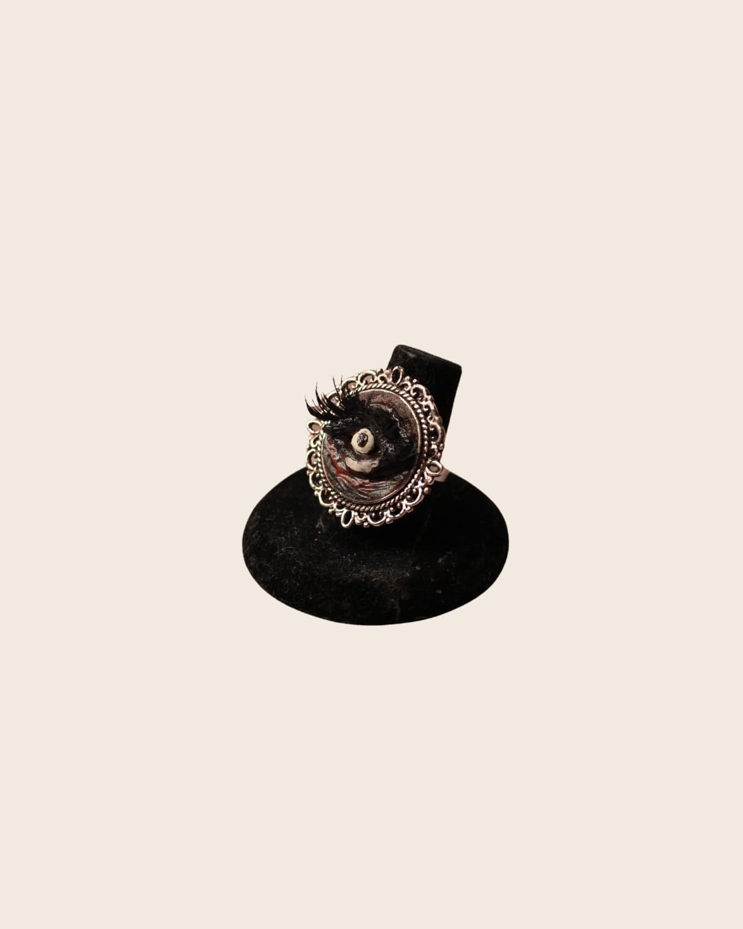 A silver ring with an ornate filigree oval setting holding a sculpted eye with black lashes and swirled dark paint, displayed on a black velvet ring stand.
