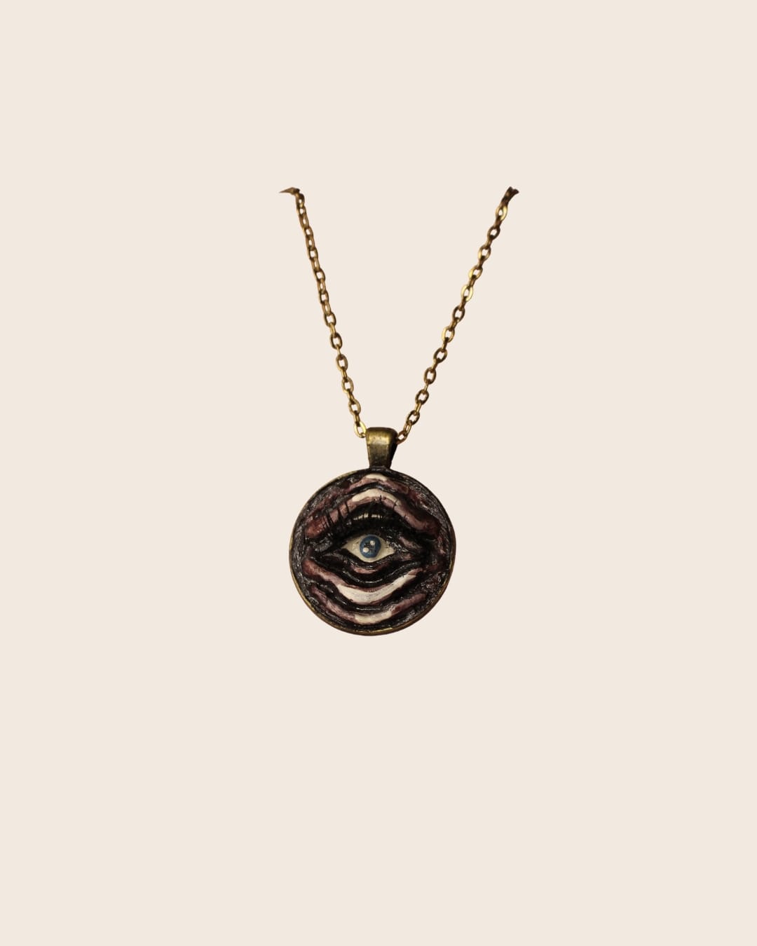 A round bronze pendant on a bronze chain, featuring a hand-sculpted blue eye with dark lashes, framed by wavy layered ridges painted in mauve, white and black.
