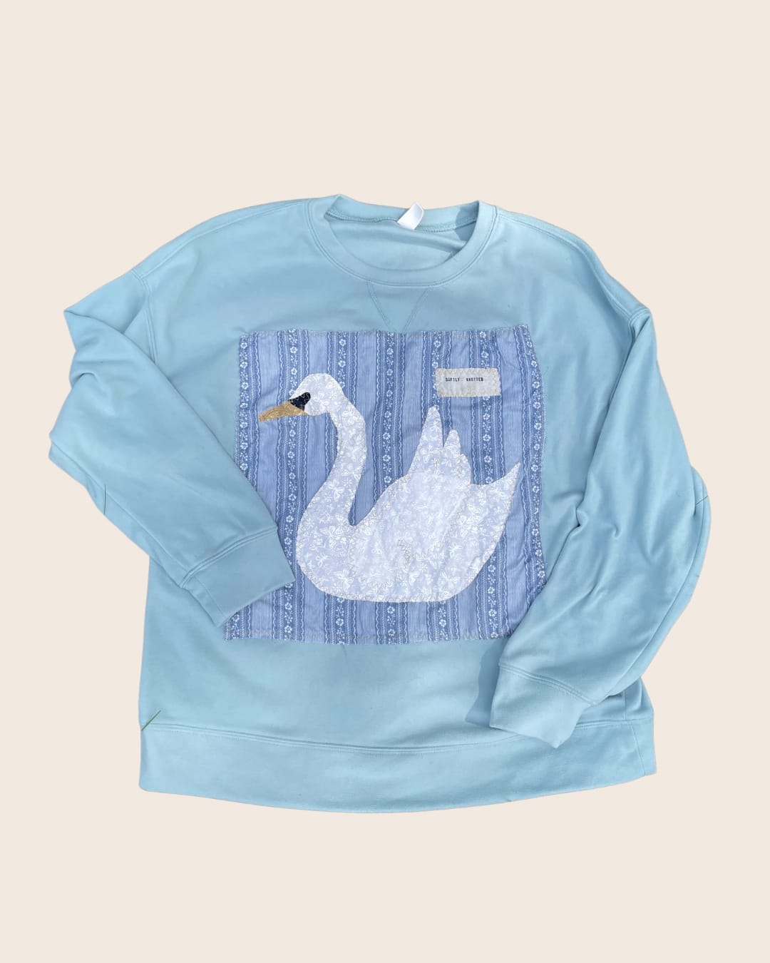 A light blue crewneck sweatshirt with a hand-sewn appliqué of a white swan on blue floral-striped fabric, with a small “Softly Knotted” label stitched in the corner.