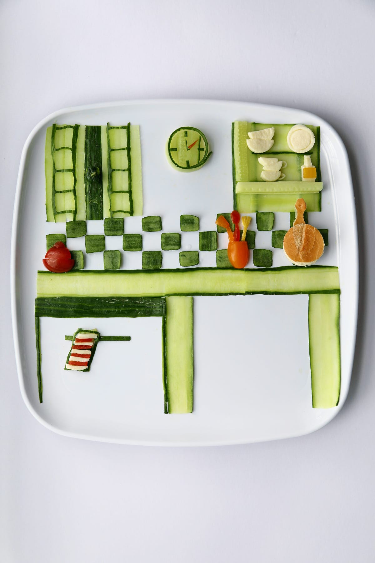 Food art on a white rectangular plate depicting a kitchen made primarily from cucumber slices, with green cabinets, a countertop, a round cucumber clock, a small stove area and tiny vegetable cooking tools.