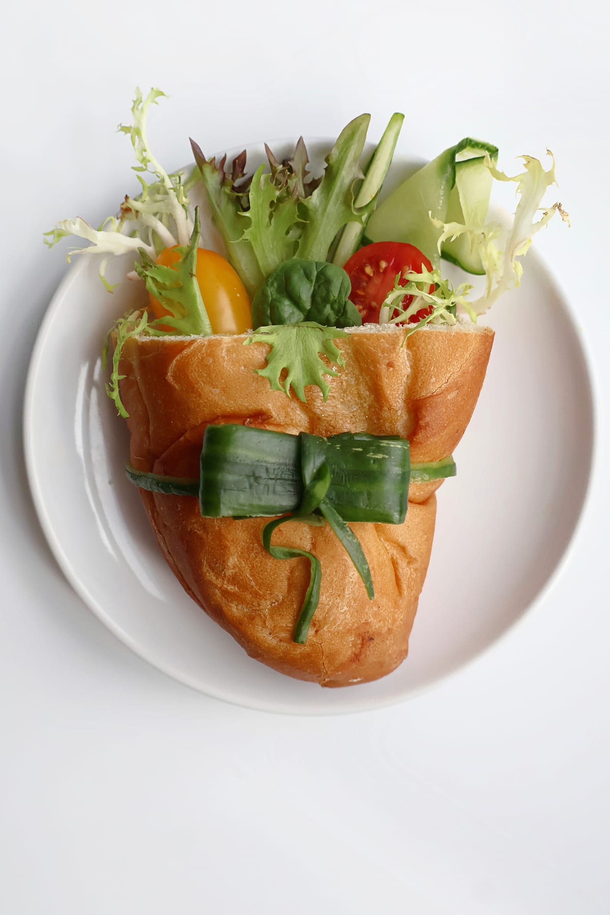 A bread pocket arranged like a bouquet on a white plate, filled with leafy greens, cucumber and red and yellow tomatoes, with a cucumber ribbon tied around the bread.