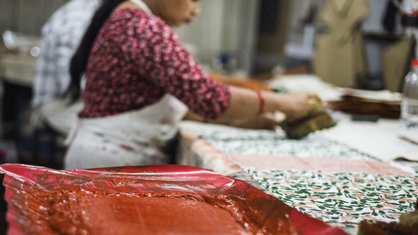 A woman in a patterned red shirt and apron block prints fabric with intricate green designs, using a red dye. The setting is a busy, artistic workspace.