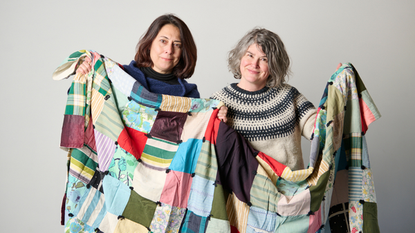 Two women facing the camera holding up a patchwork quilt.