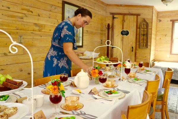 A woman setting a dining table in a rustic wood-paneled room, featuring a two-tiered food tray and marigold flower decorations.