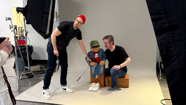 Two men pose a custom puppet dressed in a Nike jacket, cap and sneakers on a studio set surrounded by photography lights and equipment.