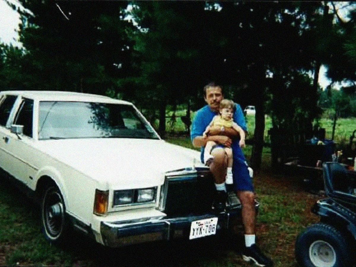 A man holding up a child in front of a white sedan.