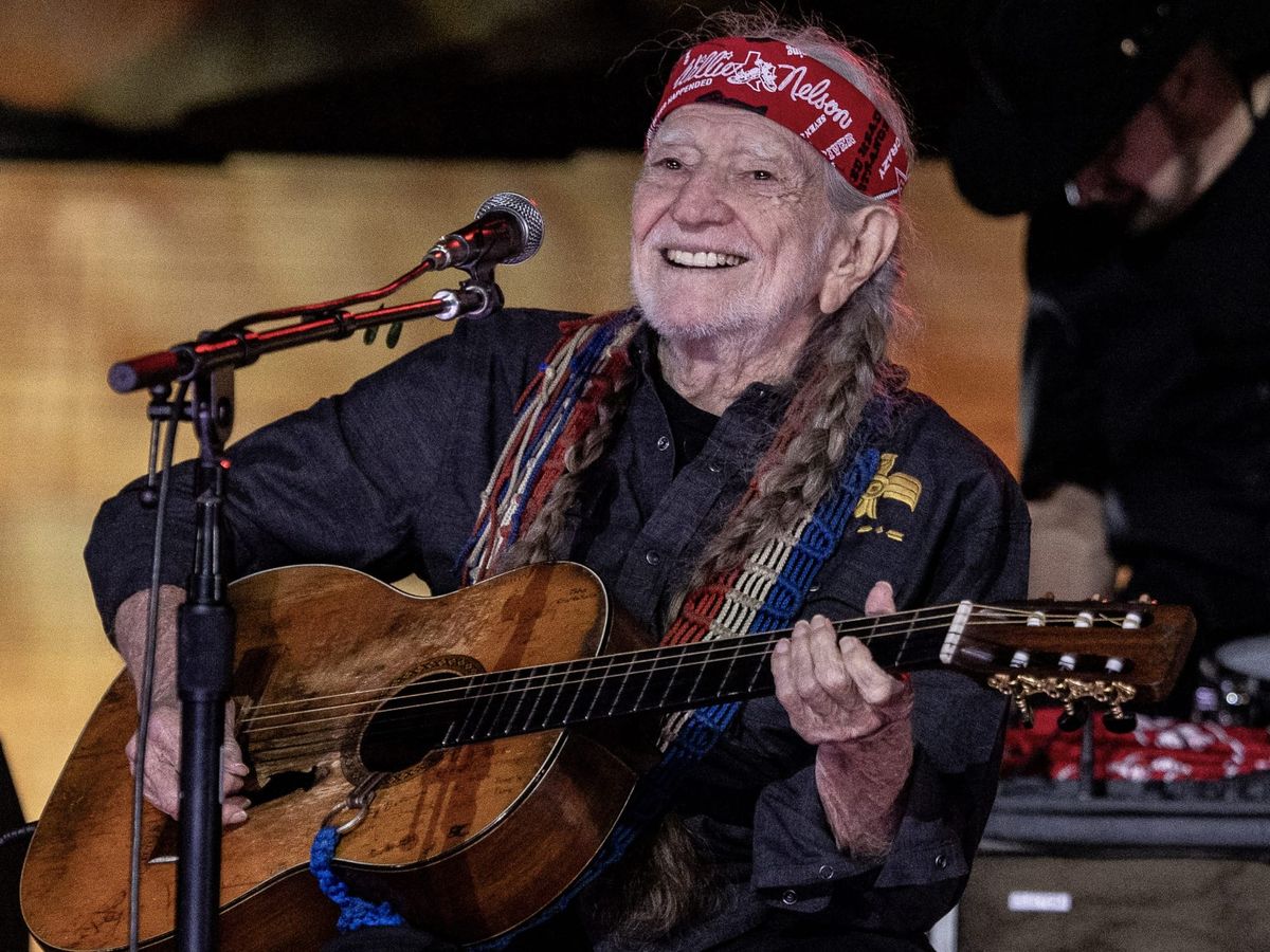 Willie Nelson on stage in a red bandana and long braids, playing guitar in front of a microphone while wearing a black long-s