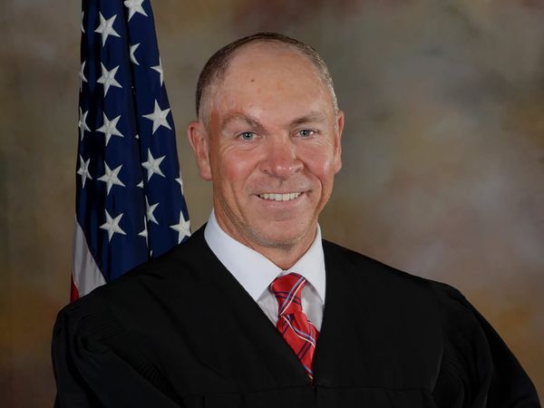 Judge Jack Pulcher smiling in front of an American flag, while wearing a robe and a red necktie