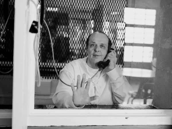 A man in prison with his hand against the glass while holding a phone