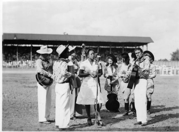 A black-and-white photo of a group of musicians dressed in white, with some wearing cowboy hats, standing in a horseshoe shap