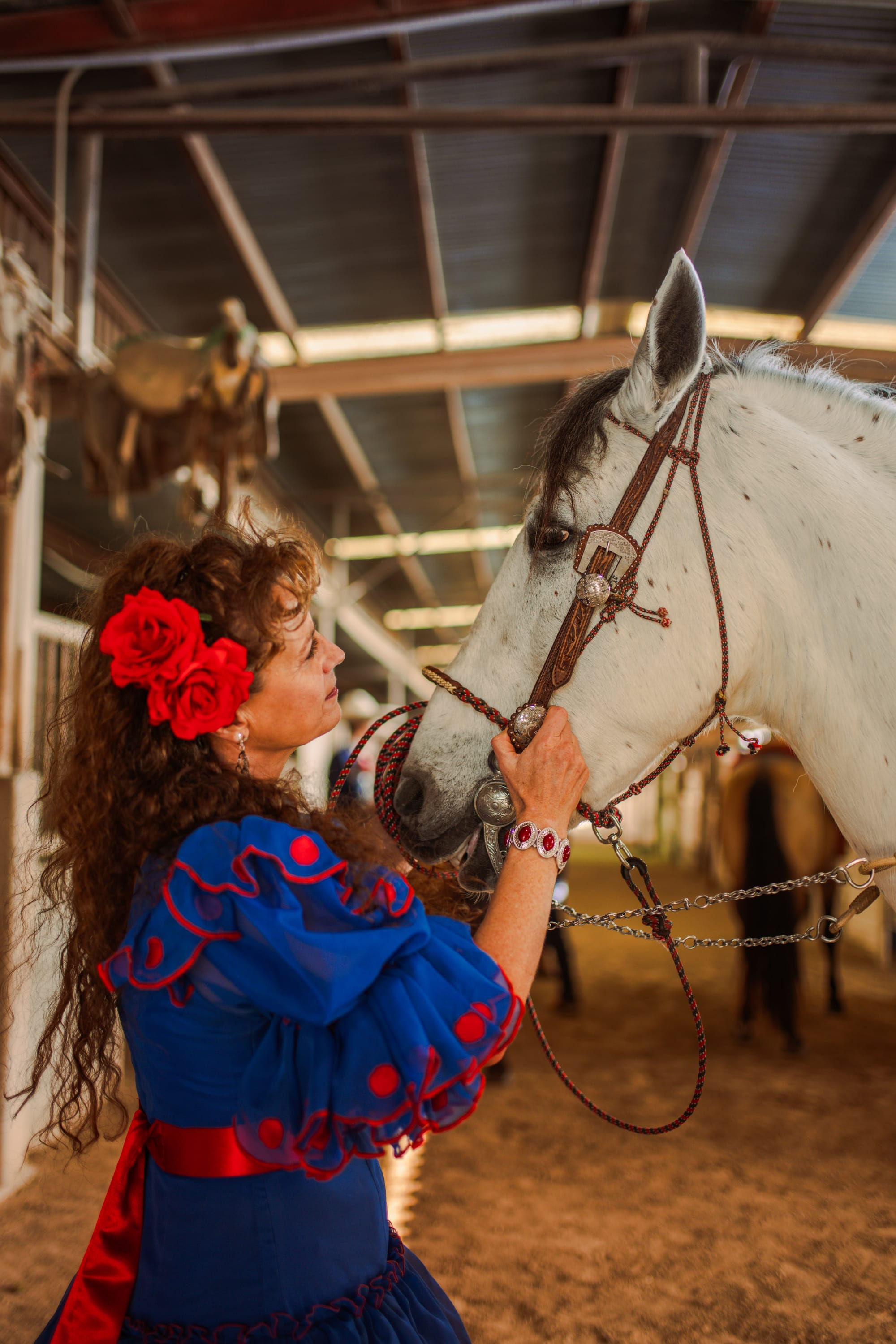 Staci, wearing red flowers in her hair and a bright blue dress, tending to a horse