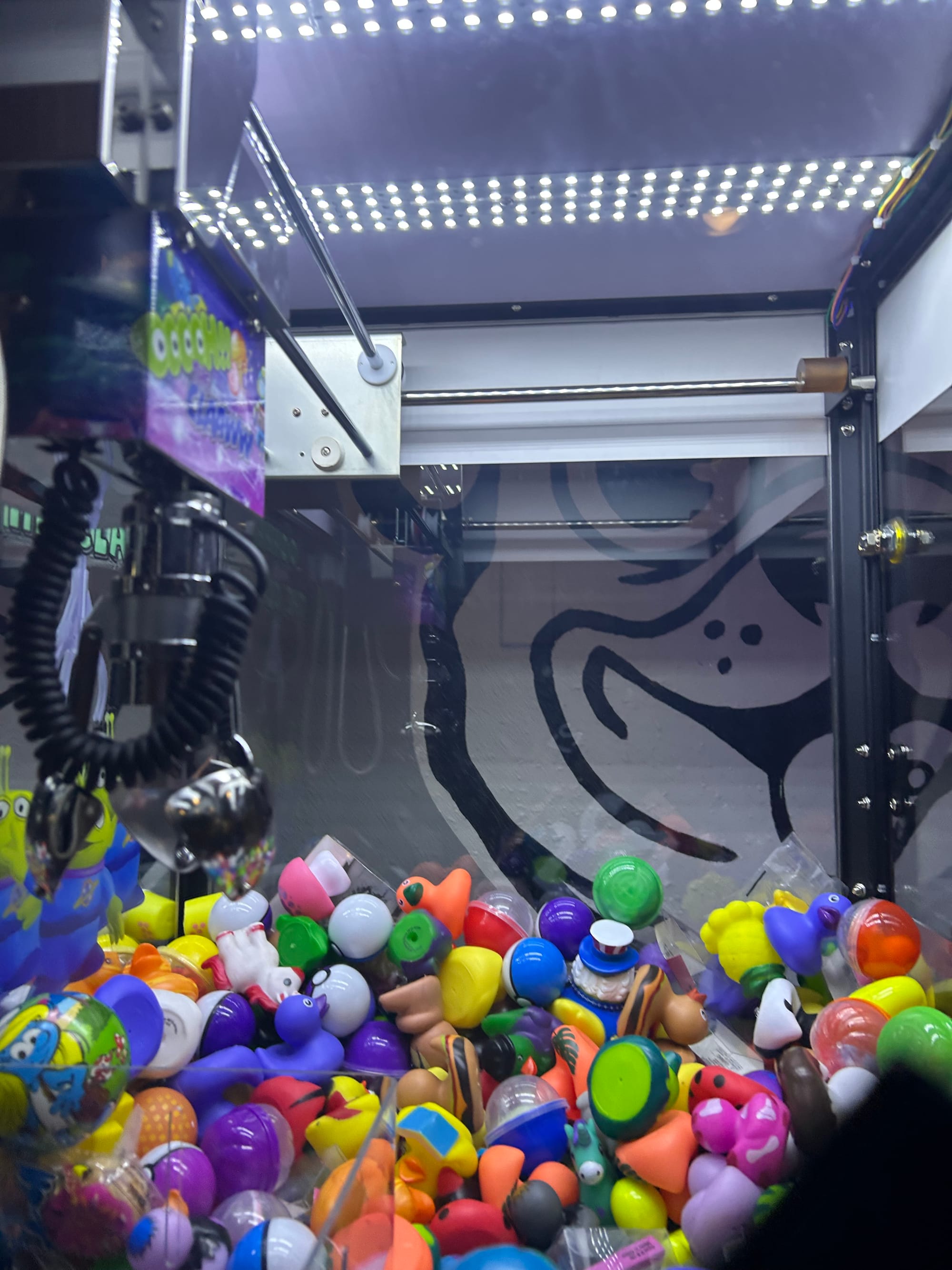 closeup of the bottom of a claw machine, full of bright plastic toys, with a partial view of a black & white mural behind it