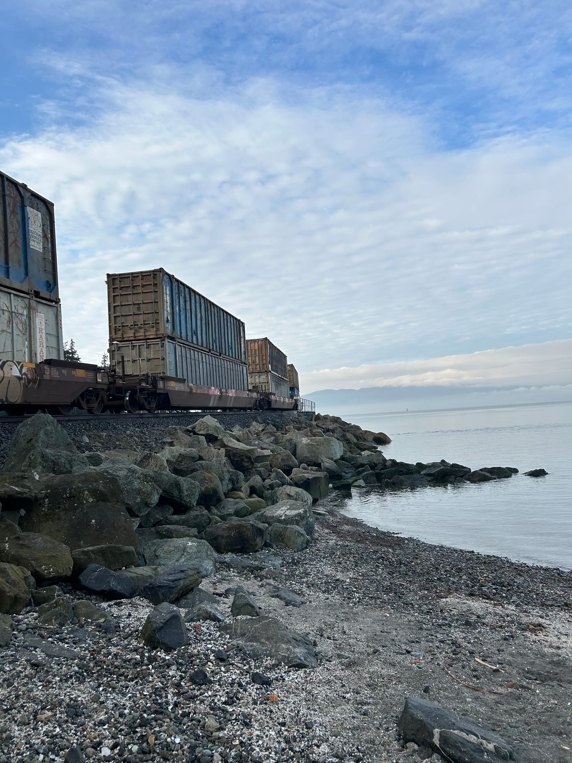 double-stacked containers on a train's curving track, sandy beach, grey-blue water, cloudy sky & islands beyond