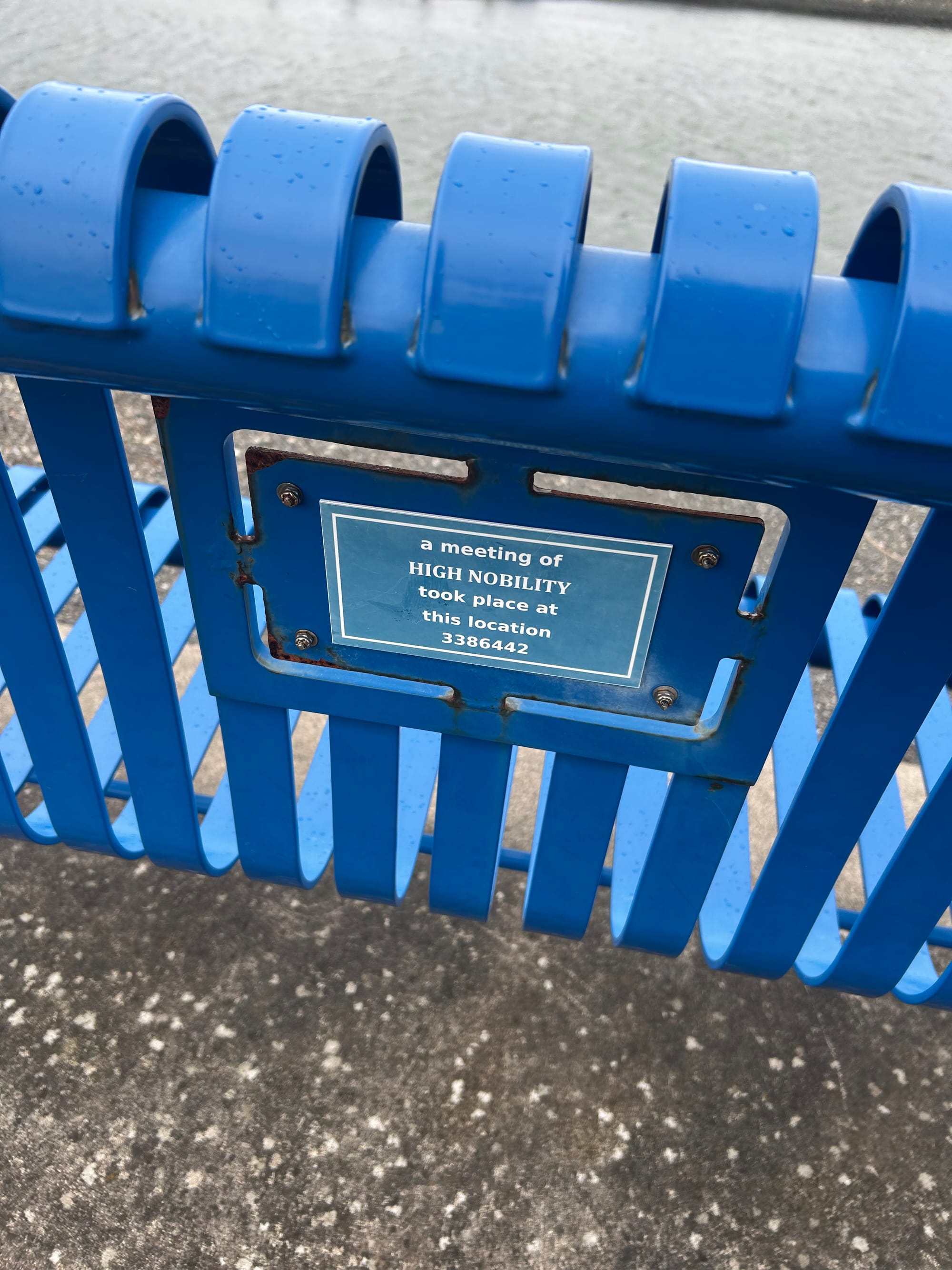 the back of a blue metal park bench, with a plaque that reads: "a meeting of HIGH NOBILITY took place at this location 3386442"