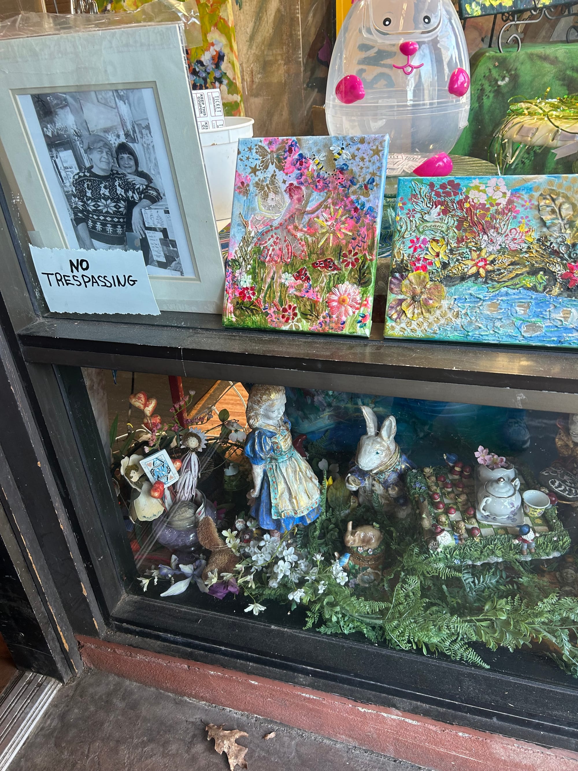 store window featuring a no trespassing sign, black & white photo of two people, small painting, a big plastic easter egg, and on the bottom a display of alice in wonderland figurines set around a tea table