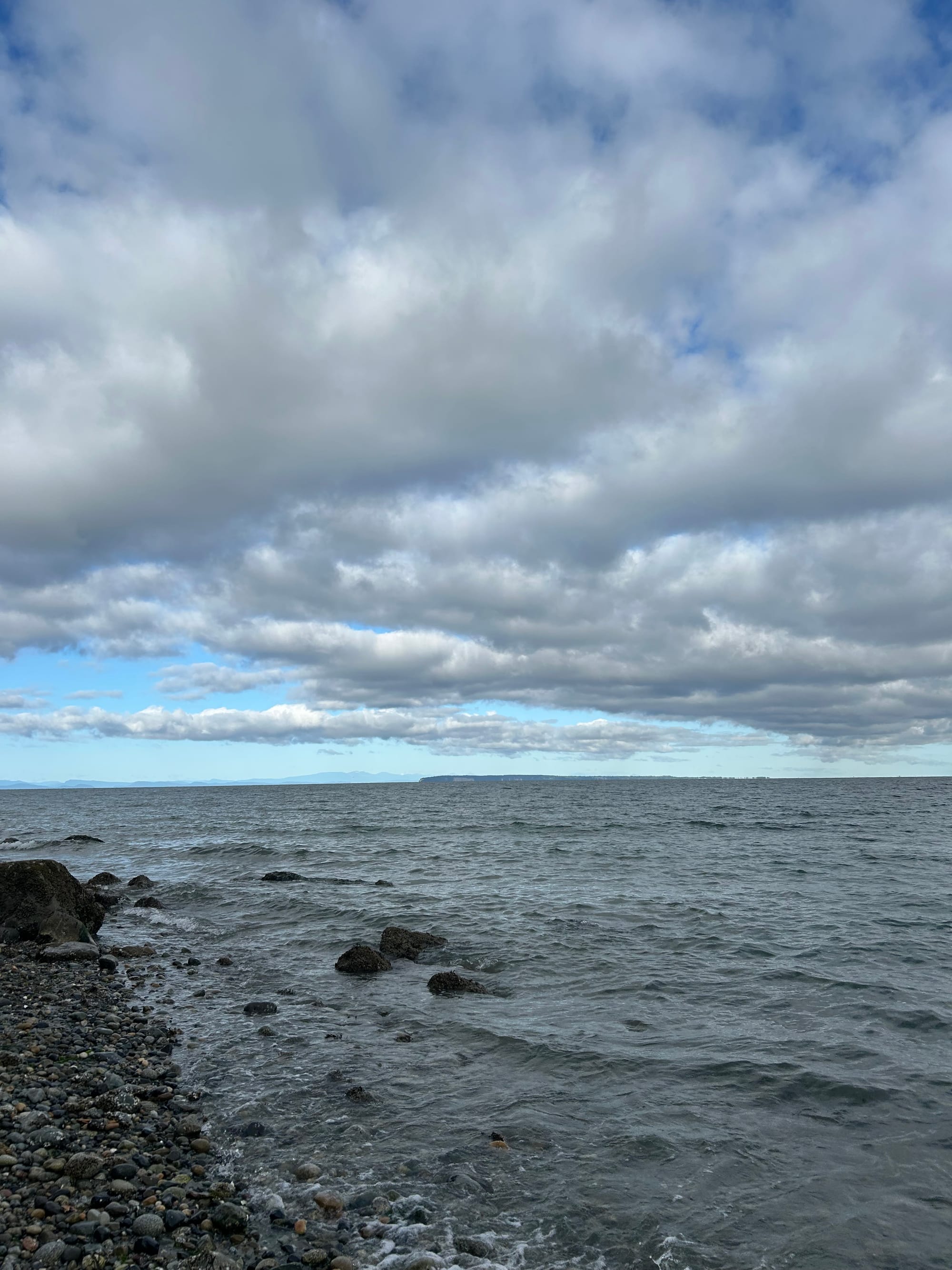 grey & white clouds overhead, streaks of blue visible, grey water with distant mountains beyond, a strip of pebbled beach 