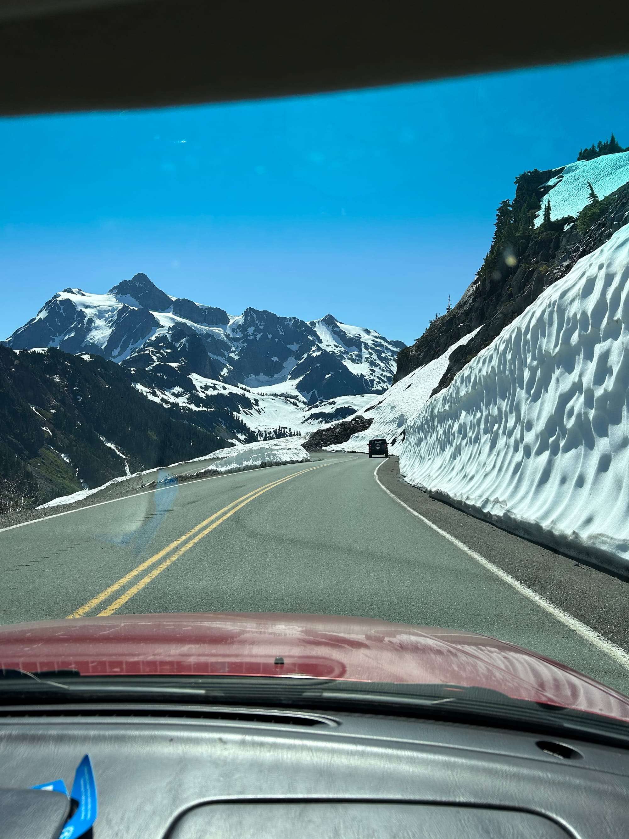 view through the windshield of eight foot high snowbanks, road curving ahead, and alpine peaks