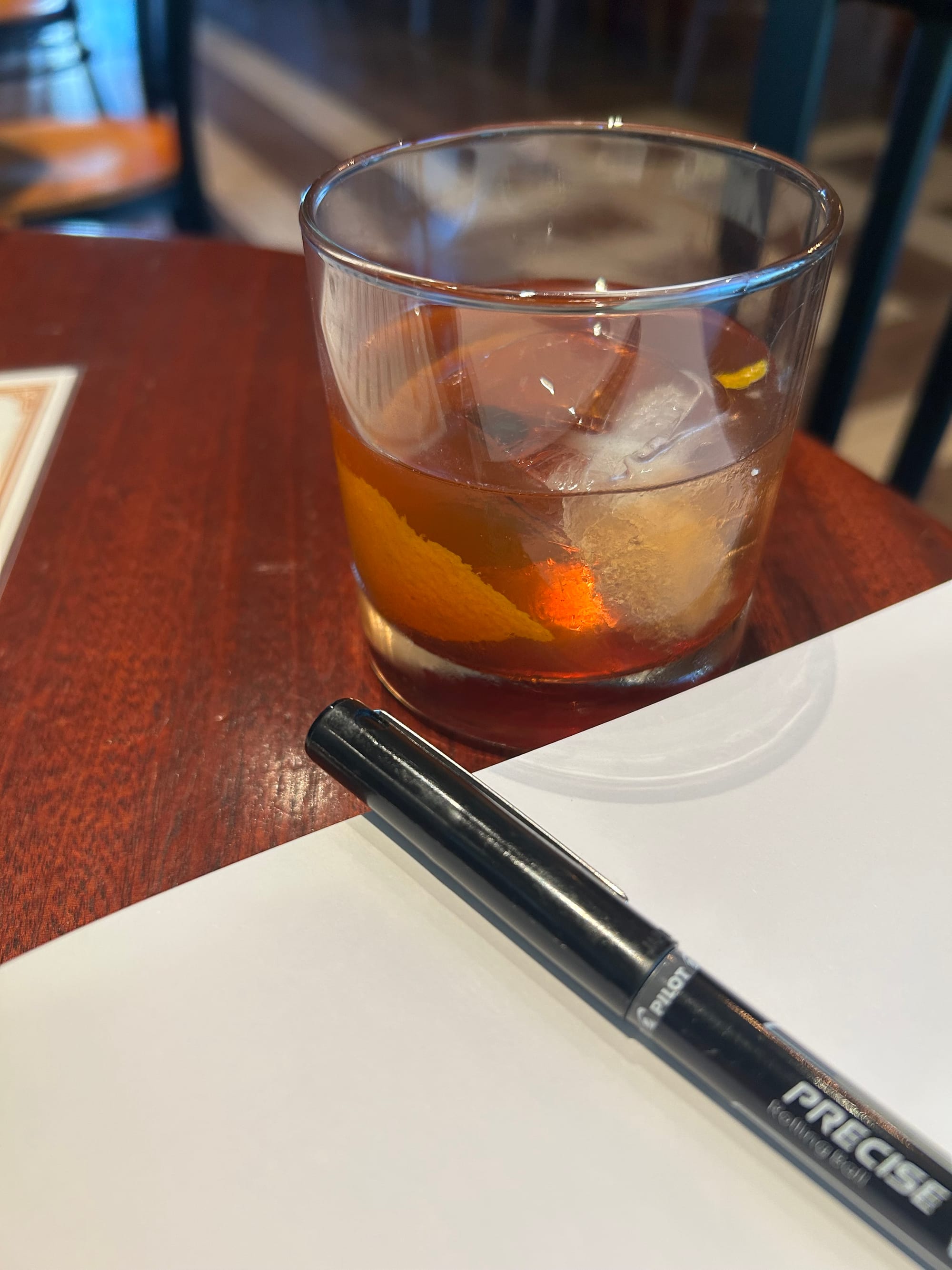 old fashioned in a glass, white notebook page, black pen, all on a wooden bar table