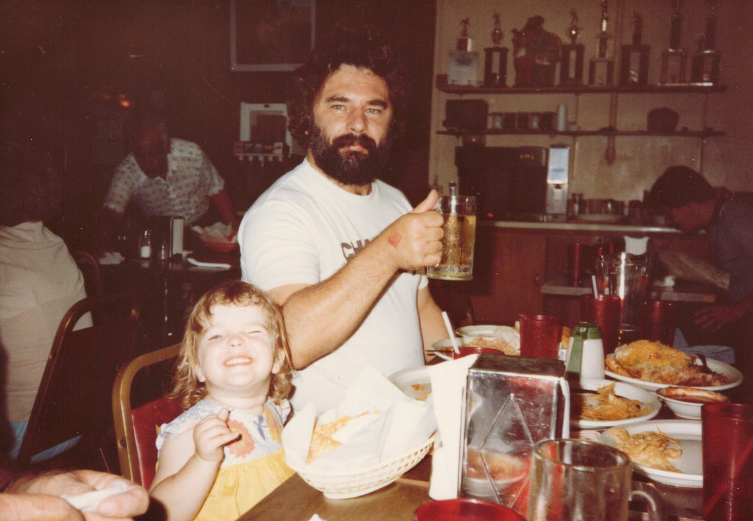 a little kid with light brown hair in a yellow top next to a bearded man in a white t shirt holding a beer, table covered in half eaten food