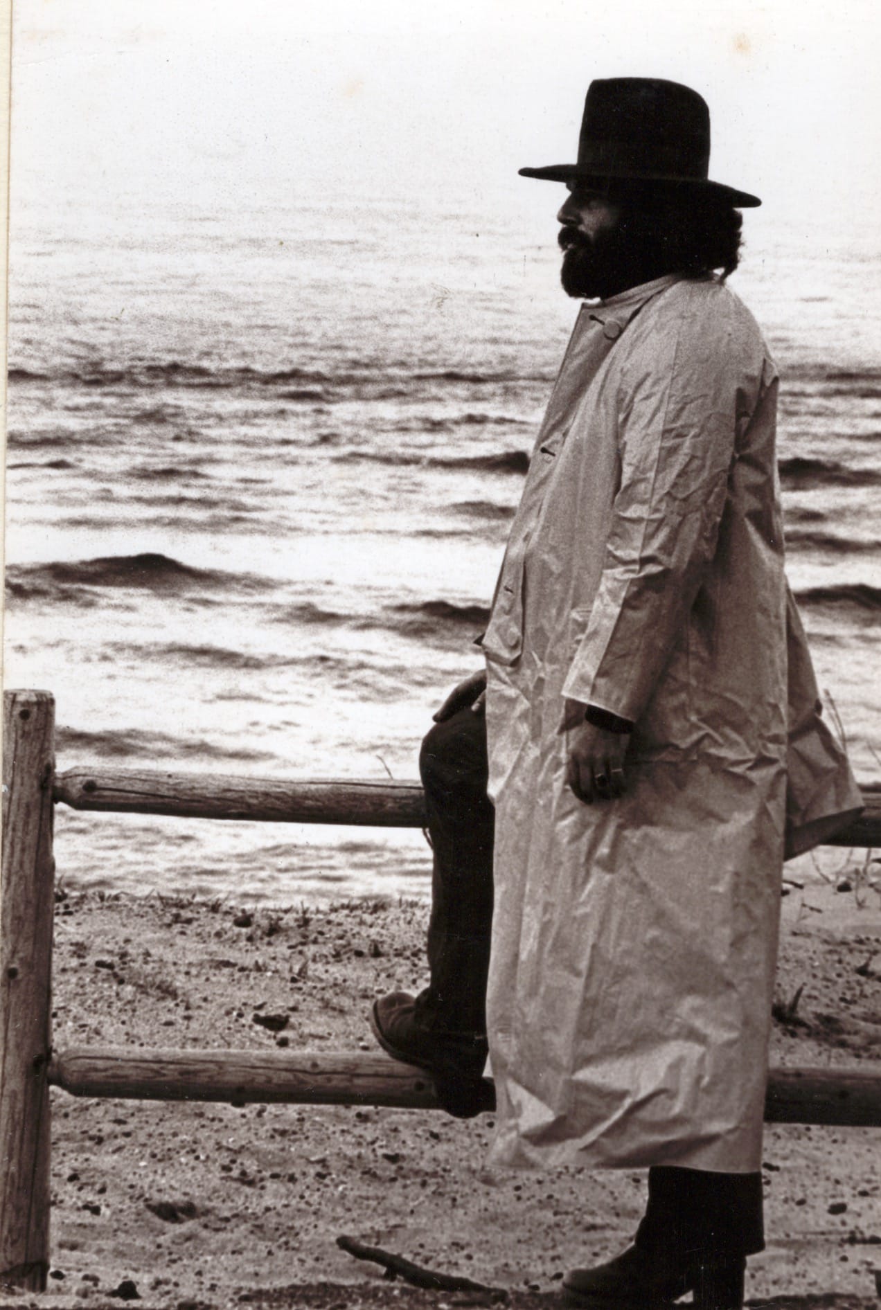 black & white photo of a tall man with a dark beard, wearing a cowboy hat, boots, & a long raincoat, standing with one foot up on a wooden fence, the ocean in the background.