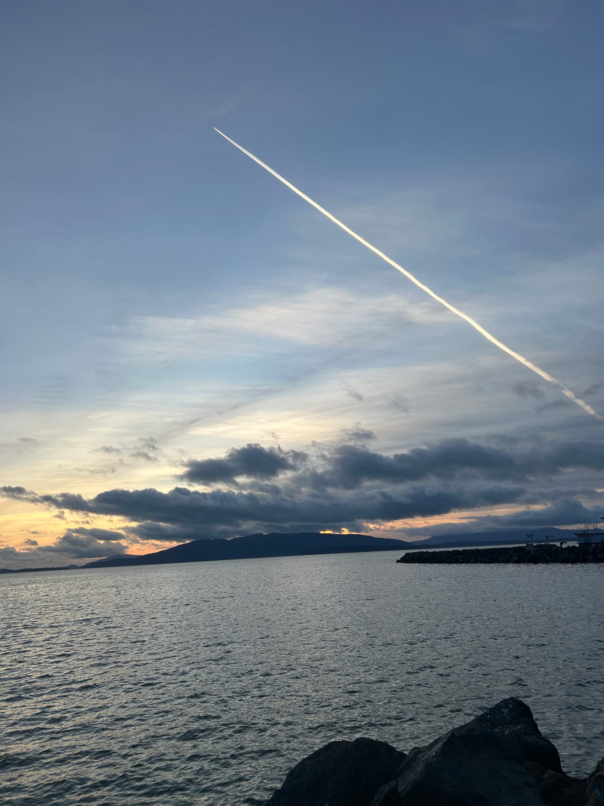 grey water, rocky outcropping, clouds & island on the horizon, long light plane trail across the sky 