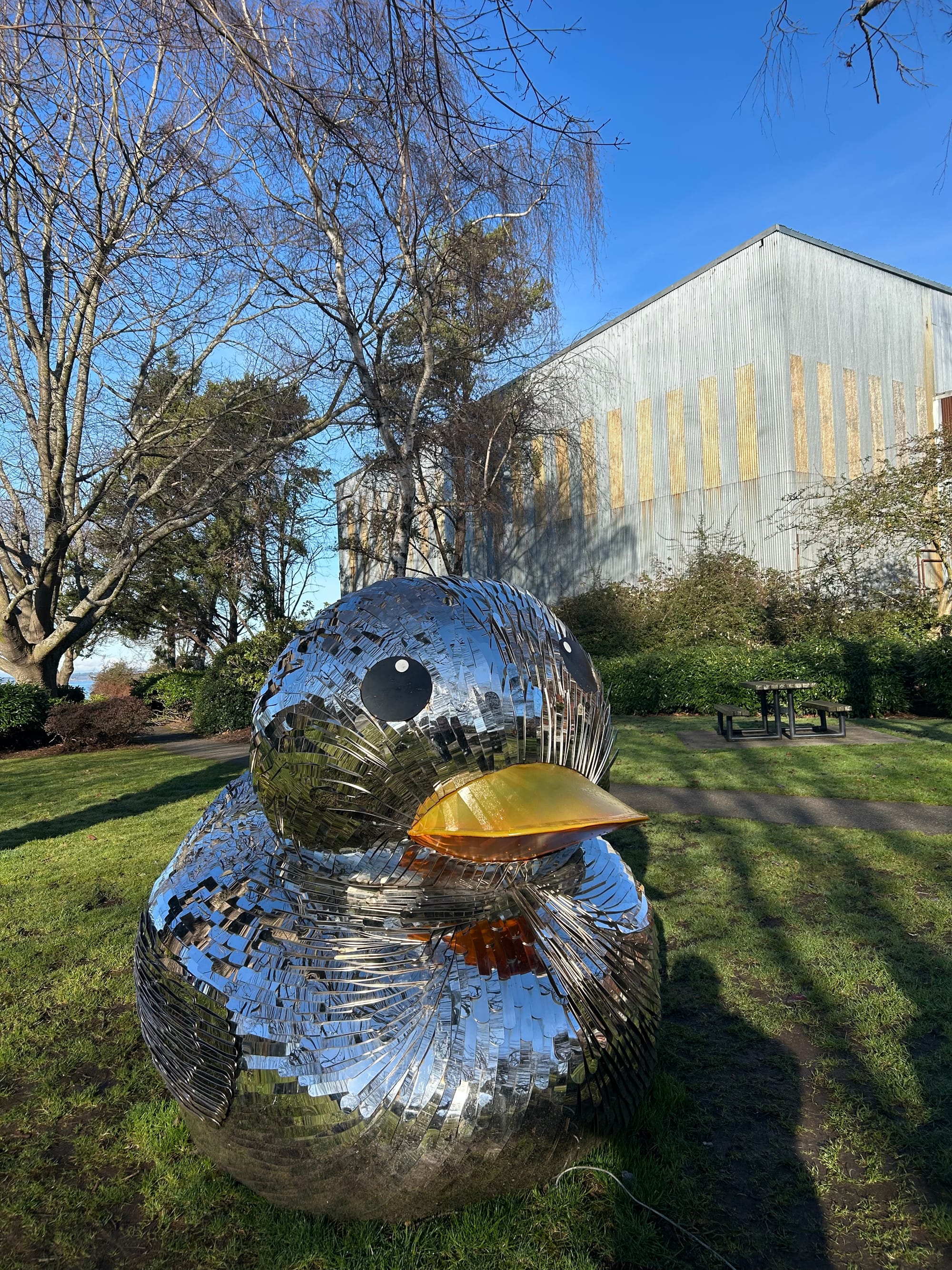 giant statue in the shape of a rubber duck with mirror pieces like a disco ball all over it & a gold beak sitting on a green lawn