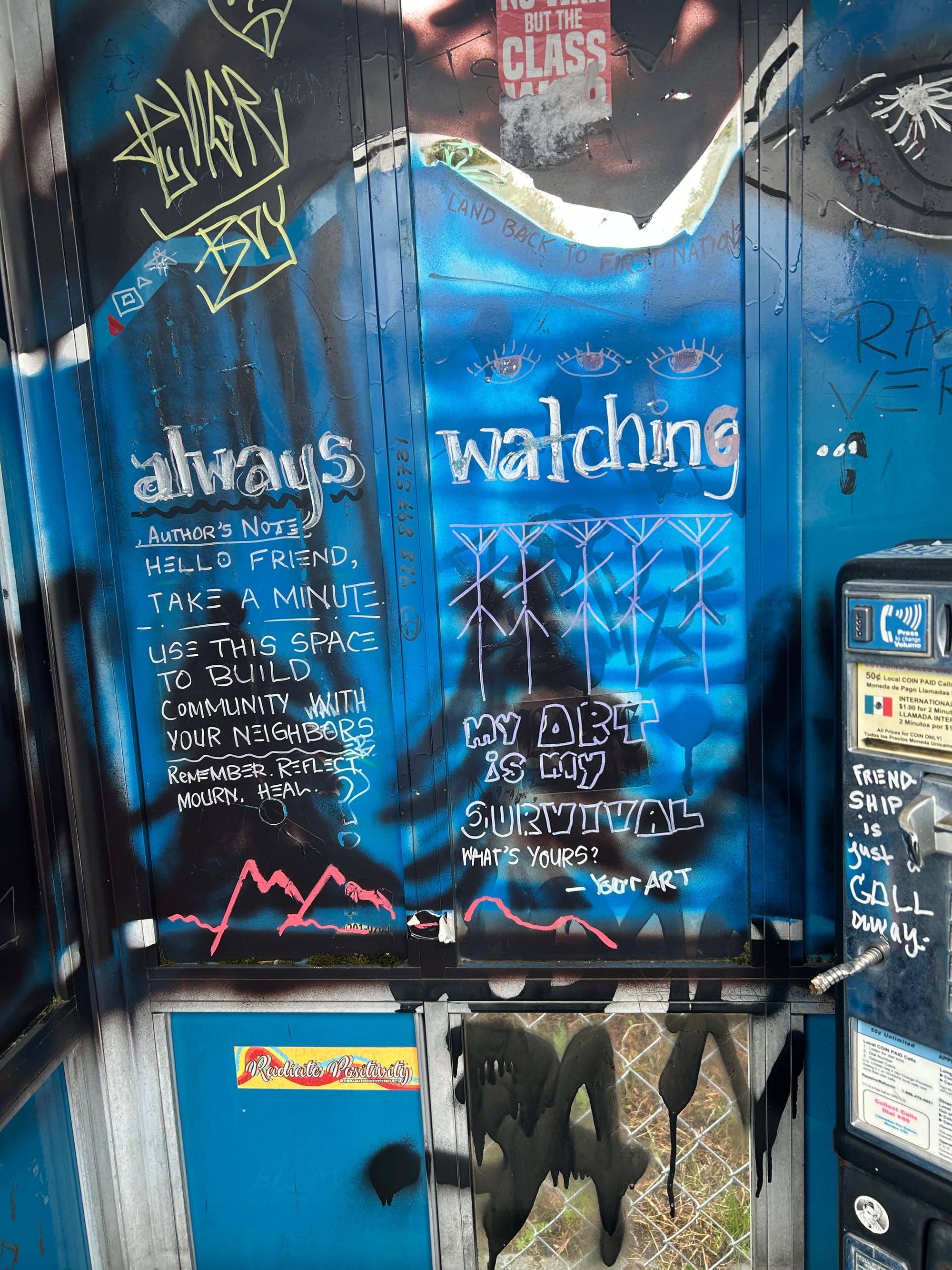 a blue phone booth graffitti'd with a number of tags, a mountain range in red, and notes that say "my art is my survival, what's yours? -- your art", "author's note: hello friend, take a minute. use this space to build community with your neighbors. remember, reflect, mourn, heal," & "always watching." on the phone itself, which has been ripped out, it says "friendship is just a call away."