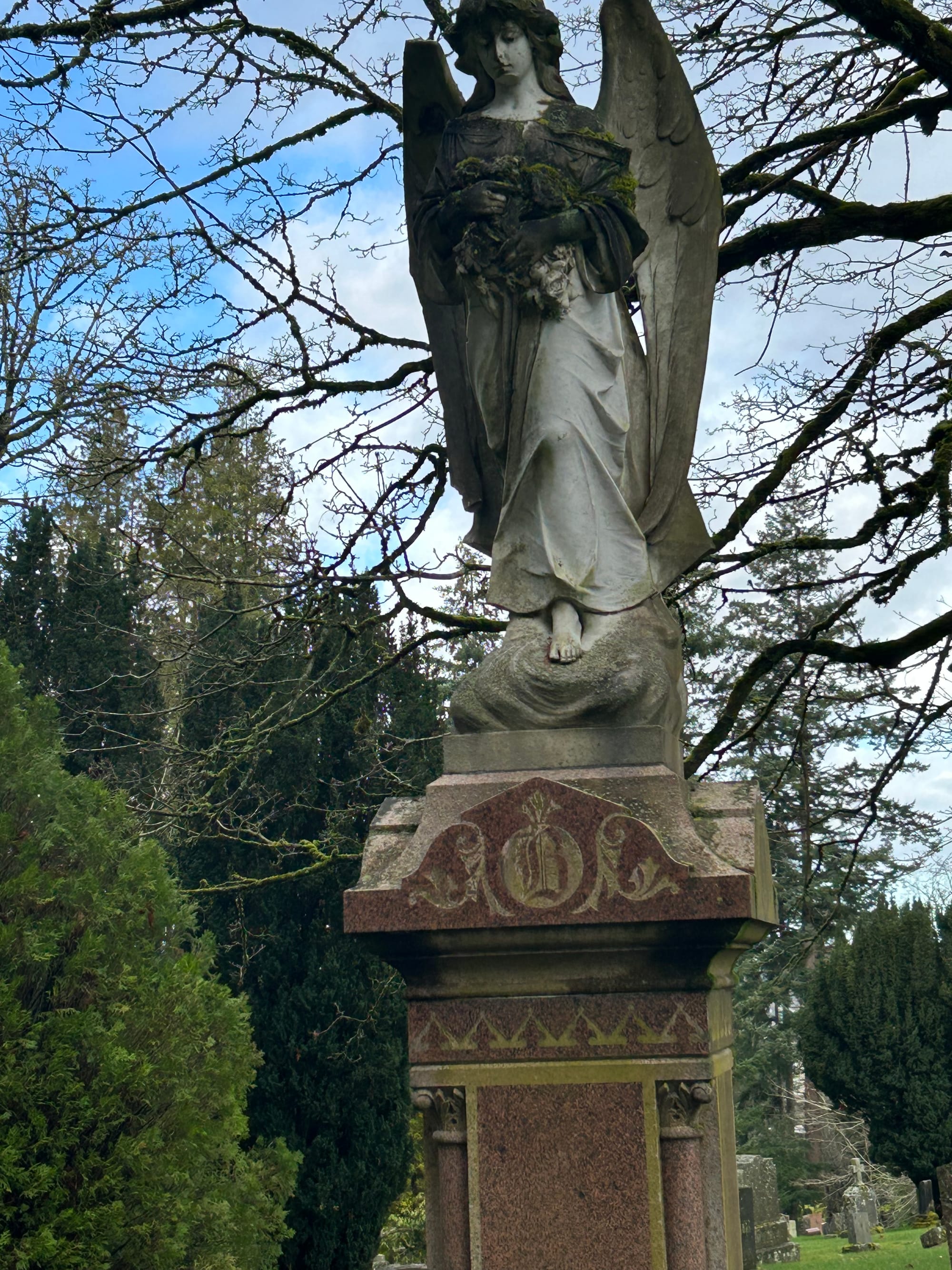 a grey statue of an angel holding a bundle of flowers, one foot extended, standing on the top of a grave. green trees and other smaller graves visible behind her.