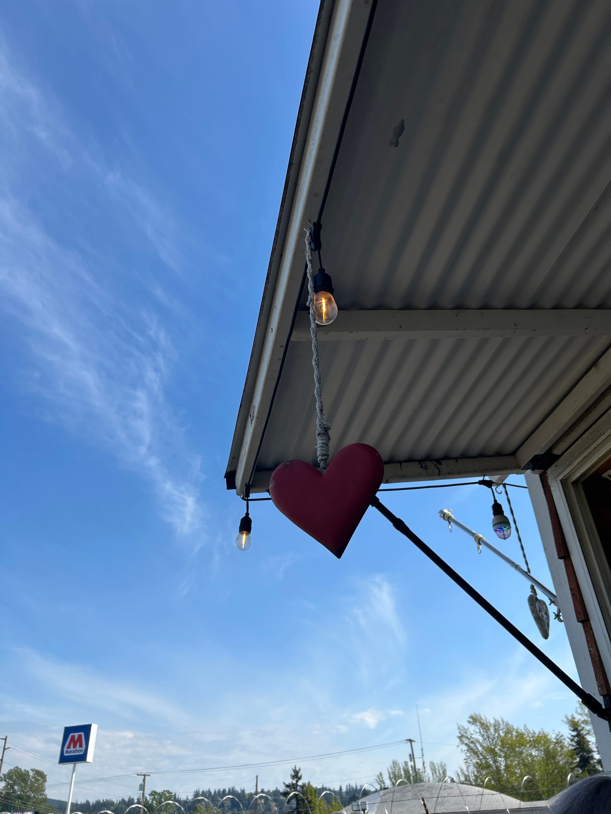 a red wooden heart dangling from a cord at the edge of a metal room, blue sky, gas station sign & barbed wire coil in the background