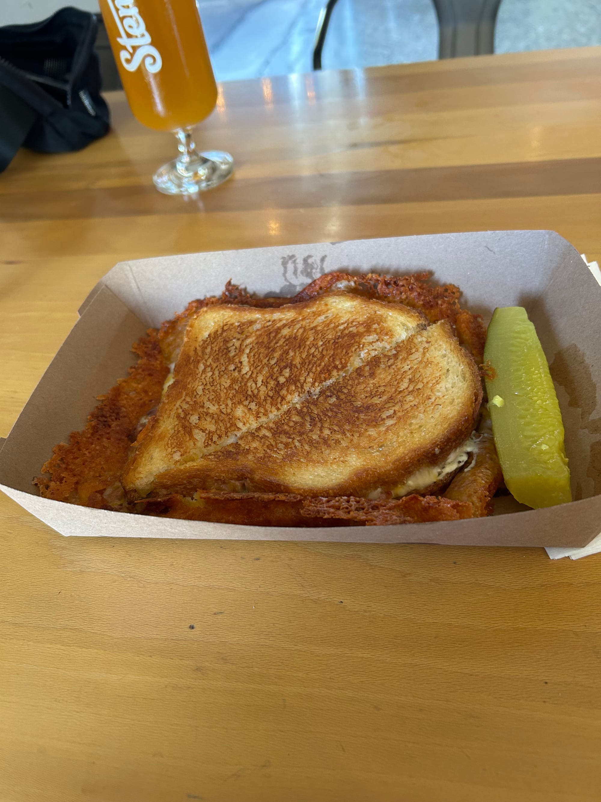 paper box with big grilled cheese sandwich, a long border of crisped cheese, and a spear of pickle