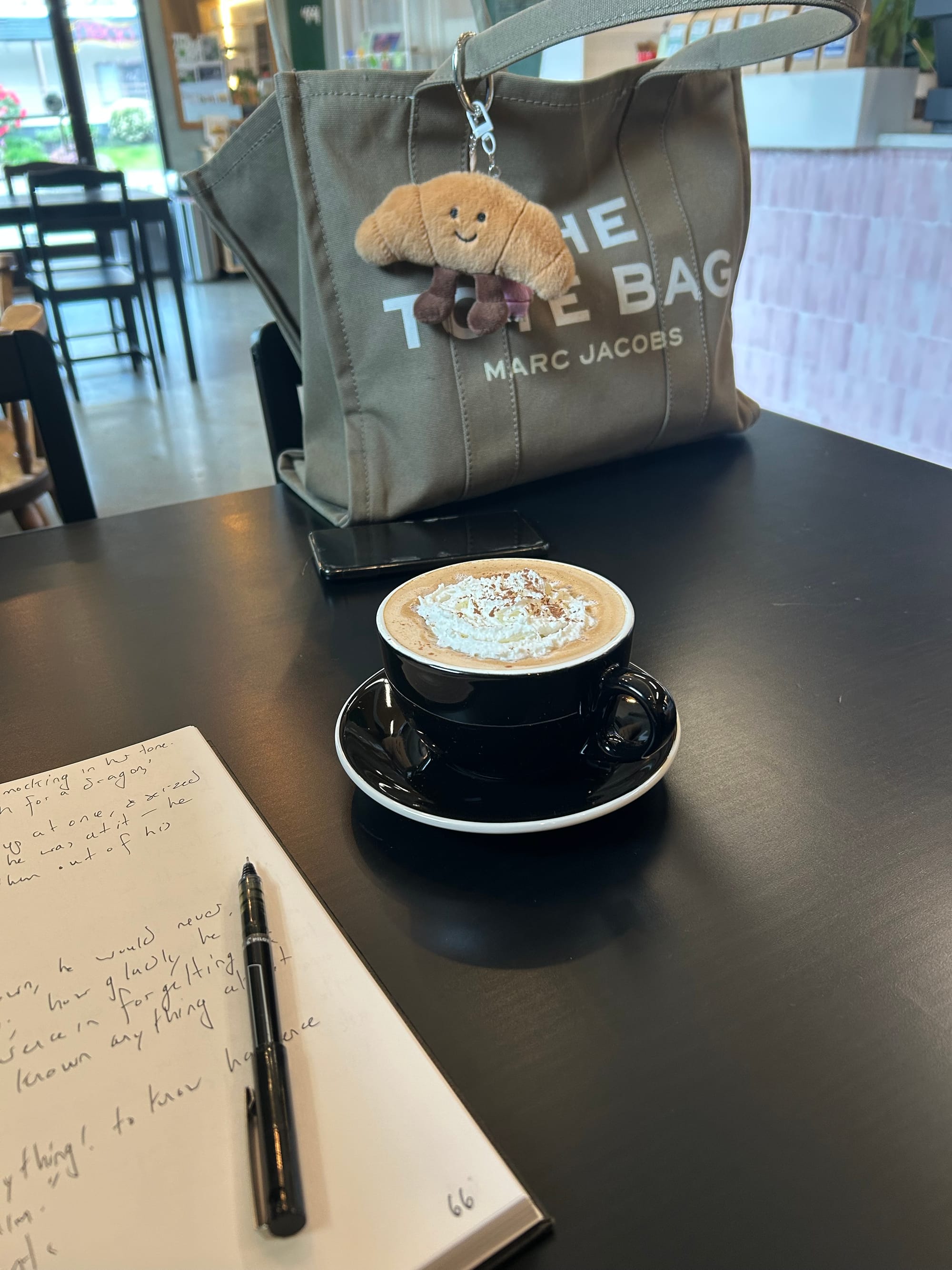 open notebook with white paper & black handwriting scribbled, a black saucer & mug with a mocha & whipped cream, grey Marc Jacobs tote bag with croissant bag charm all sitting on a black cafe table, pink tile in the background