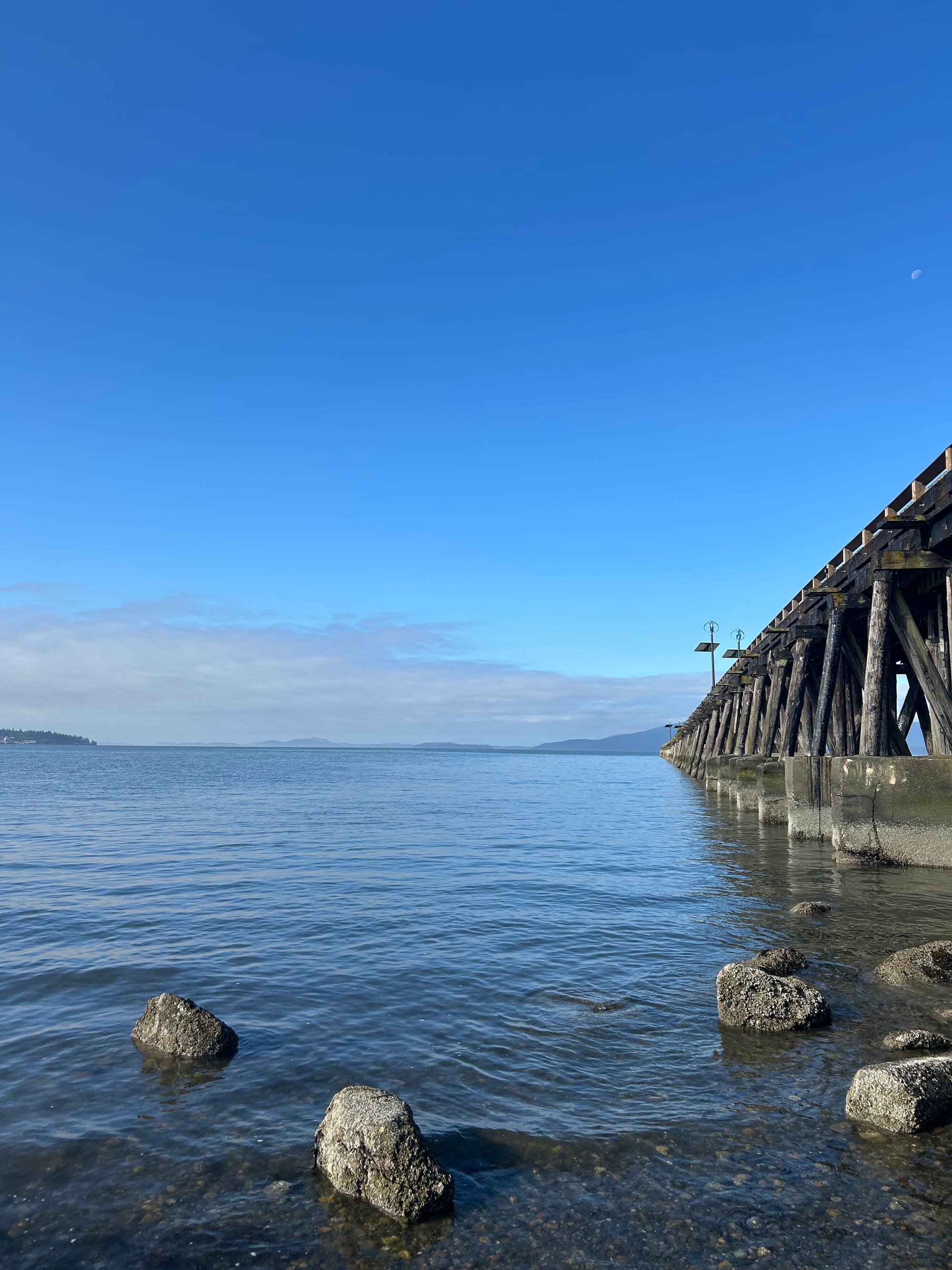 pebble beach with water right up to the frame of the picture, a few barnacle covered rocks, a long pier stretching off the the right, clouds at the horizon of a blue sky, islands in the distance