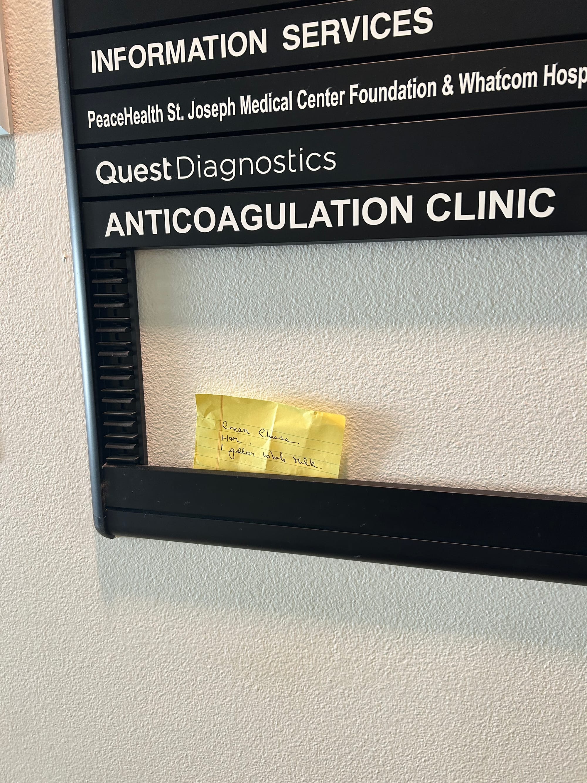 a black sign for a medical building listing occupants, into which a small scrap of yellow paper has been inserted. The yellow paper reads: "Cream chesee, ham, 1 gallon whole milk."