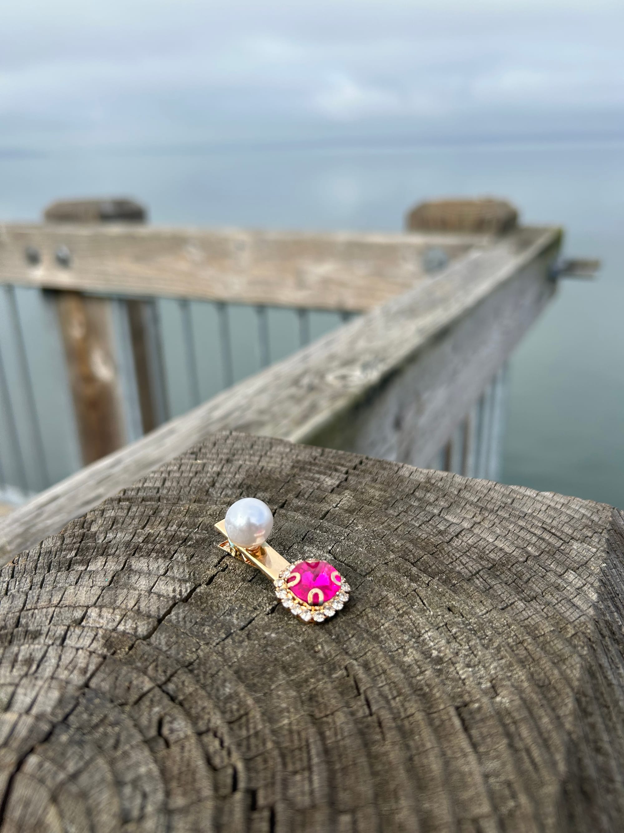 clip on costume earring, gold with pink fake gem & a pearl, set on a wooden post overlooking grey water