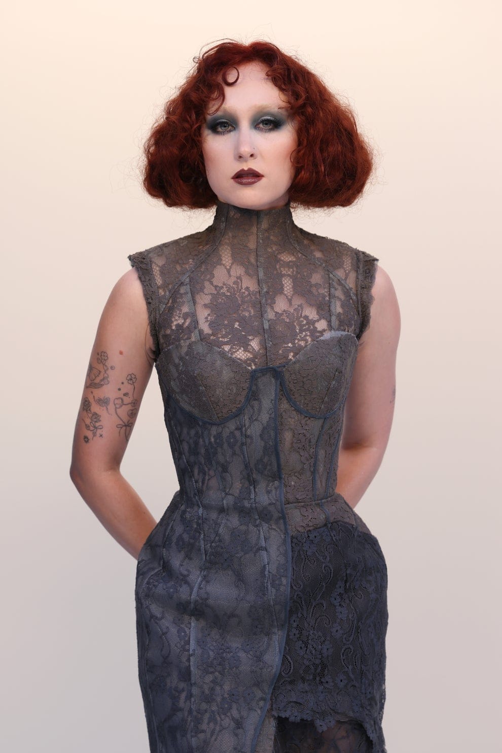 Person with curly hair on a red carpet in a sleeveless, form-fitting lace gown with tattooed arms, giving a confident expression