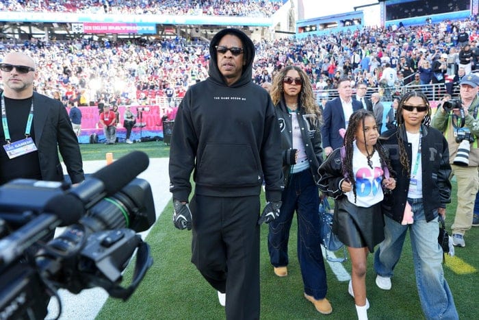A group of people, including a man in a casual hoodie and sunglasses, walk on a football field surrounded by a crowd