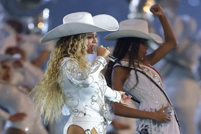 Singer performing on stage in an embellished bodysuit and wide-brimmed hat, with dancers in matching hats and outfits in the background