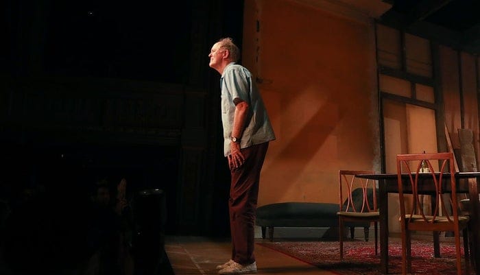 Person on stage performing, wearing a casual shirt and pants, with a dramatic set backdrop featuring chairs and a table