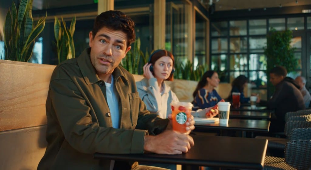 Adrien Grenier in a green jacket holding a Starbucks drink while sitting at a table with other people.
